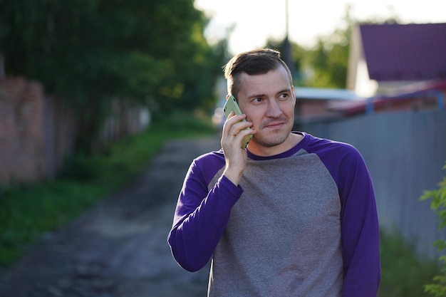 Hombre hablando por teléfono inteligente en un suburbio Hombre adulto con ropa informal respondiendo llamadas telefónicas y mirando hacia otro lado mientras está parado en el fondo borroso de una calle suburbana