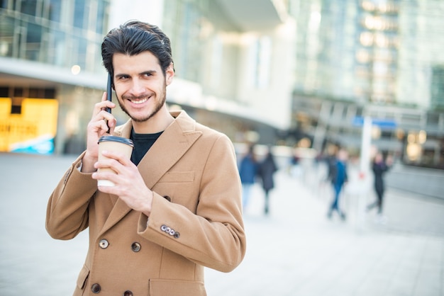 Hombre hablando por teléfono y sosteniendo una taza de café mientras camina en una ciudad