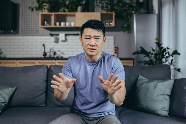 Hombre hablando por webcam enojado gesticulando con las manos asiáticas mirando la cámara llamada en línea sentado en el sofá en casa