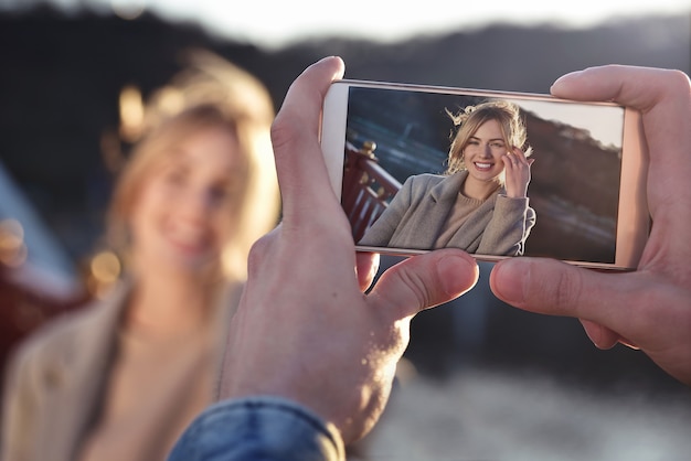 Un hombre hace un retrato en el teléfono de la mujer sonriente feliz de pie en el puente en el soleado día de primavera o otoño al aire libre