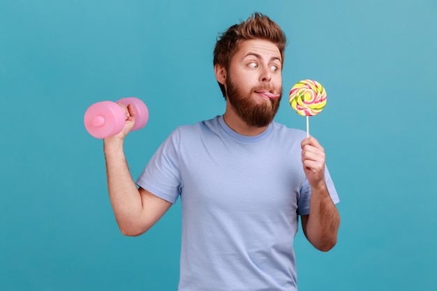 Hombre haciendo ejercicio con mancuerna rosa mirando sabroso lollypop con la lengua fuera quiere morder dulces