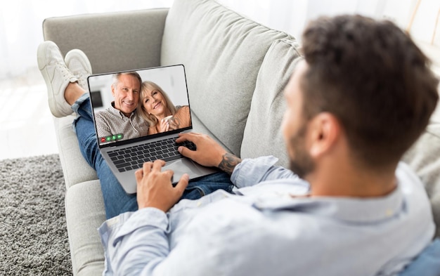 Hombre haciendo videollamadas en una computadora portátil con padres mayores mientras se relaja en el sofá