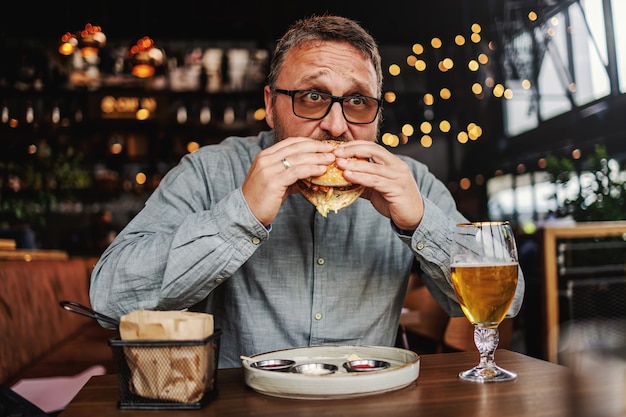 Hombre hambriento barbudo de mediana edad sentado en el restaurante y comiendo una deliciosa hamburguesa.