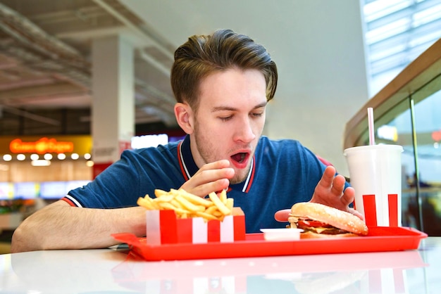 El hombre hambriento está sentado en el patio de comidas comiendo una hamburguesa grande y sabrosa Comida chatarra gorda rápida Hambre