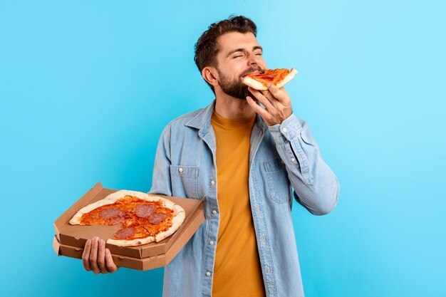 Foto hombre hambriento del milenio disfruta del sabor de la pizza con telón de fondo azul