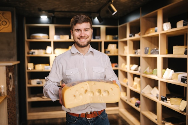 Hombre hansome sostiene una gran rebanada de queso maasdam en la mano Queso con grandes agujeros Fondo de estantes con queso