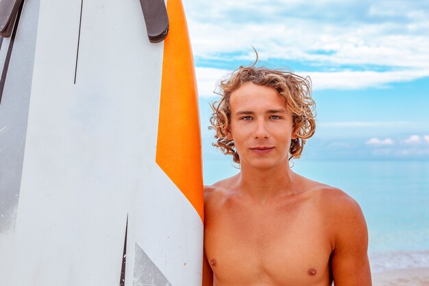 El hombre hermoso camina con la tabla de surf en blanco blanca espera la onda para surfear el lugar en la orilla del mar del océano. Concepto de deporte, fitness, libertad, felicidad, nueva vida moderna, hipster.