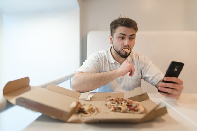 Un hombre hermoso y divertido come una pizza de la caja y levanta su teléfono. Selfies con pizza