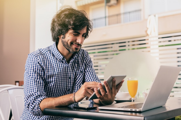 Hombre hermoso joven en el café, sosteniendo una tableta y trabajando en línea con su computadora portátil