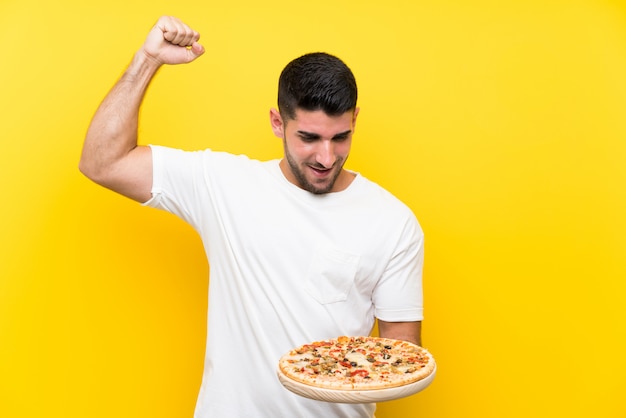 Hombre hermoso joven que sostiene una pizza sobre la pared amarilla aislada que celebra una victoria