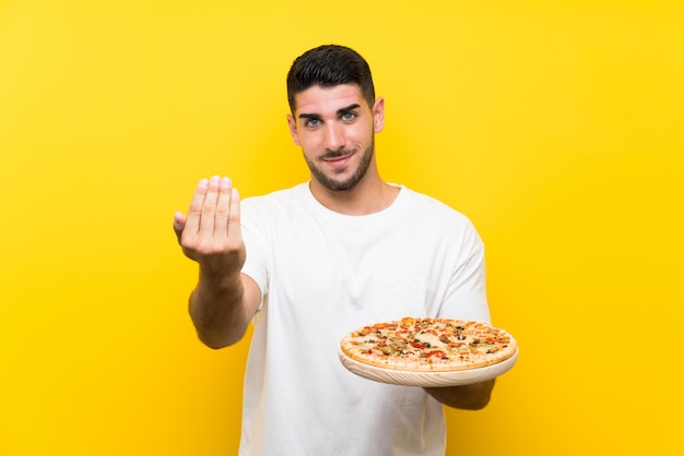 Hombre hermoso joven que sostiene una pizza sobre la pared amarilla aislada que invita a venir con la mano