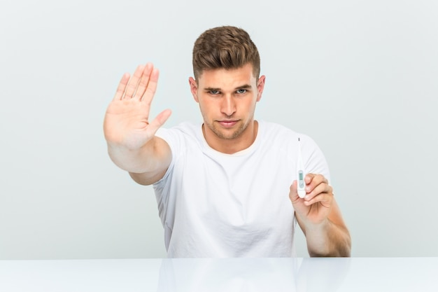 Hombre hermoso joven que sostiene un termómetro que se coloca con la mano extendida que muestra la señal de stop, previniéndole.