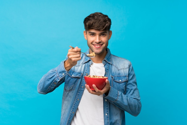 Hombre hermoso joven sobre la pared azul aislada que sostiene un tazón de cereales