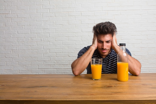 Hombre hermoso y natural joven que se sienta en una tabla frustrada y desesperada, enojada y triste con las manos en la cabeza. Desayunando, incluye zumo de naranja y un bol de cereales.