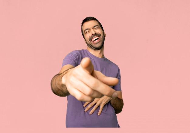 Hombre hermoso que señala con el dedo en alguien y que ríe mucho en fondo rosado aislado