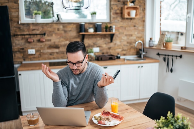 Hombre hermoso que tiene problemas que compran en línea con una tarjeta de crédito y un ordenador portátil por la mañana en la tabla de la cocina con el desayuno.