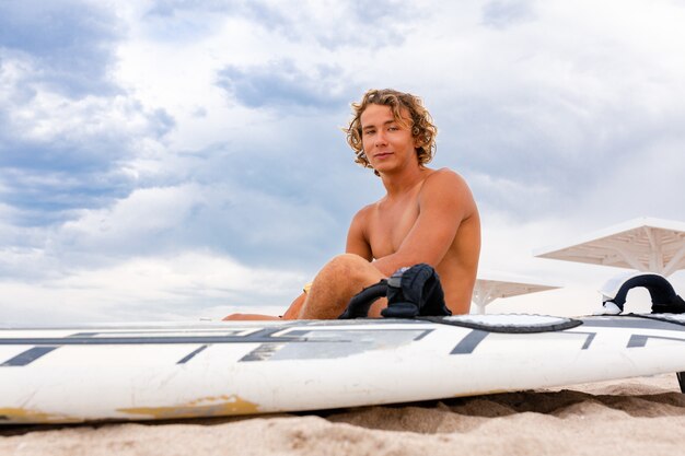 El hombre hermoso se sienta en la playa con la tabla de surf blanca en blanco espera la onda para surfear el lugar en la orilla del mar.