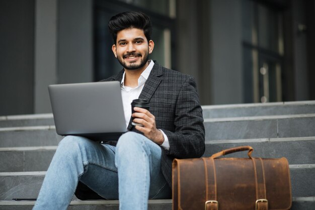 Hombre hindú sonriente usando una computadora portátil mientras está sentado en las escaleras