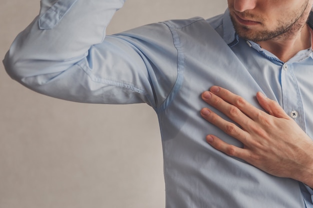 Hombre con hiperhidrosis sudando mucho debajo de la axila con camisa azul, sobre gris.