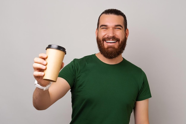 El hombre hipster con una amplia sonrisa te está dando una taza de café para llevar fresca