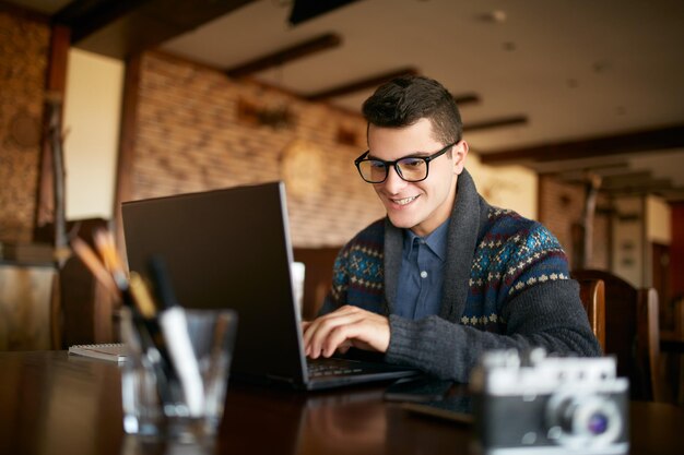 Hombre hipster atractivo y sonriente con gafas y suéter de invierno trabajando en una computadora portátil en un café Empresario independiente navegando por Internet en una cafetería Fotógrafo retocando fotos Cámara vintage en la mesa