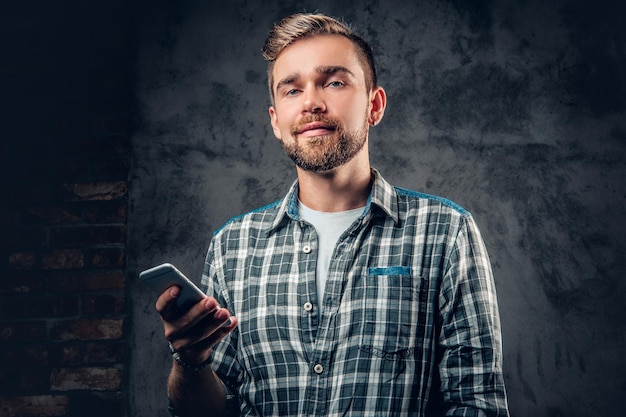 Hombre hipster barbudo en una camisa de lana sostiene un teléfono inteligente sobre fondo gris.