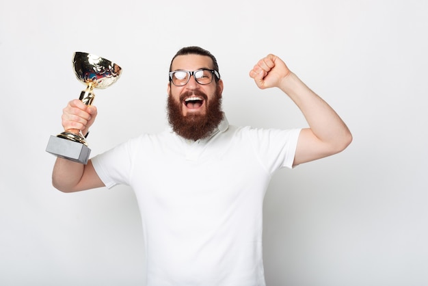 Hombre hipster barbudo sorprendido en camiseta blanca celebrando y sosteniendo la copa de campeón