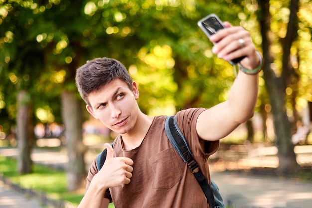 Hombre hipster en camiseta marrón guiña un ojo y muestra clase a la cámara del teléfono en el parque de la ciudad verde de verano