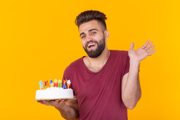 Hombre hipster joven guapo positivo en camiseta burdeos con pastel de felicitación con inscripción feliz cumpleaños posando en una pared amarilla. Concepto de felicitaciones y aniversarios.