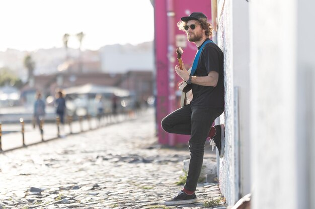 Un hombre hipster parado junto a la pared y tocando la guitarra eléctrica en la calle