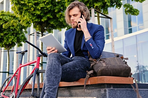 Hombre hipster con pelo largo y rubio habla por teléfono inteligente y tiene tablet PC con bicicleta de una sola velocidad en el fondo.