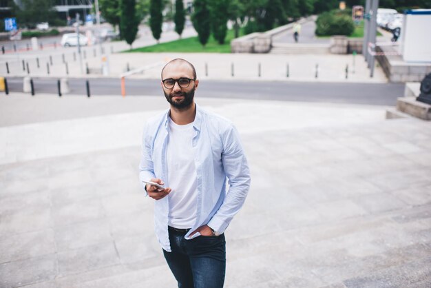 Hombre hipster positivo usando teléfono inteligente mientras camina por la calle urbana