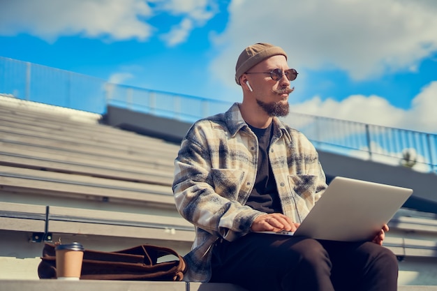 Hombre hipster se sienta al aire libre con café mientras trabaja blogs estudiando en línea mirando hacia los lados