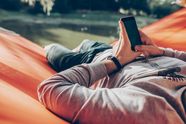 Hombre hipster sosteniendo un teléfono inteligente con pantalla vacía con espacio para texto y relajándose en una hamaca en un parque soleado de verano tipo simulacro mirando el concepto de redes sociales de teléfono en blanco