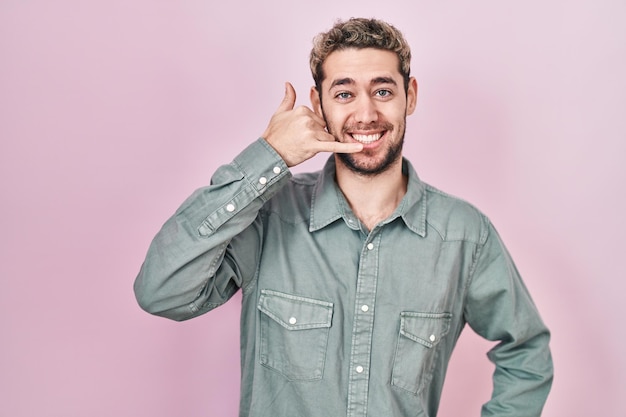 Hombre hispano con barba sobre fondo rosa sonriendo haciendo gestos telefónicos con la mano y los dedos como hablar por teléfono. comunicar conceptos.