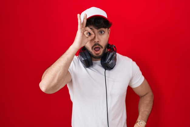 Hombre hispano con barba con sombrero de jugador y auriculares haciendo un gesto correcto sorprendido con la cara sorprendida mirando a través de los dedos expresión incrédula