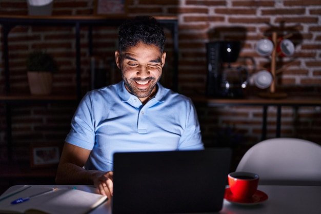 Hombre hispano con barba usando laptop en la noche guiñando un ojo mirando a la cámara con expresión sexy, cara alegre y feliz.