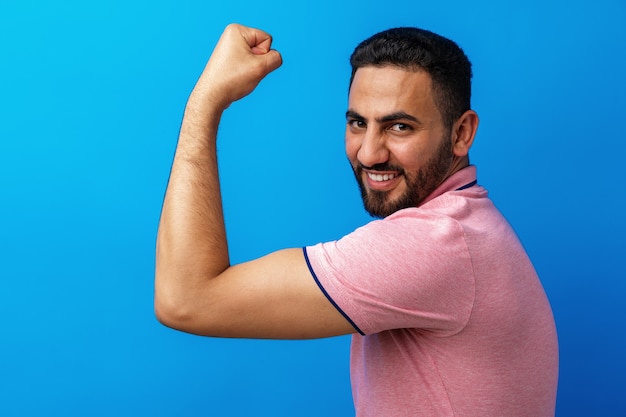 Hombre hispano con barba vistiendo camiseta casual mostrando los músculos de los brazos contra el fondo azul.