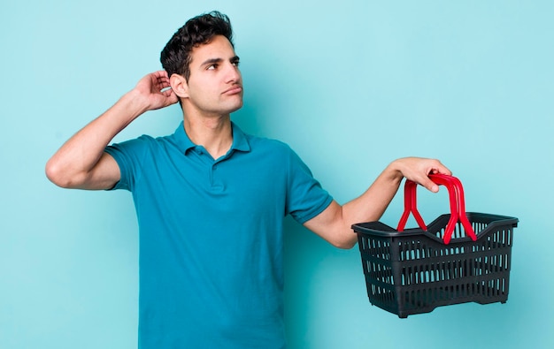Hombre hispano guapo que se siente perplejo y confundido rascándose la cabeza concepto de cesta de la compra