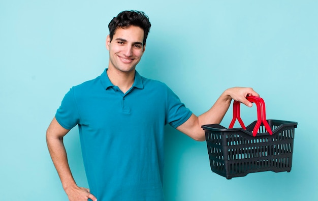 Hombre hispano guapo sonriendo felizmente con una mano en la cadera y un concepto de cesta de la compra seguro