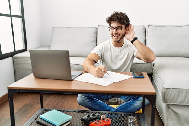 Hombre hispano haciendo papeles en casa sonriendo con la mano sobre la oreja escuchando rumores o chismes. concepto de sordera.