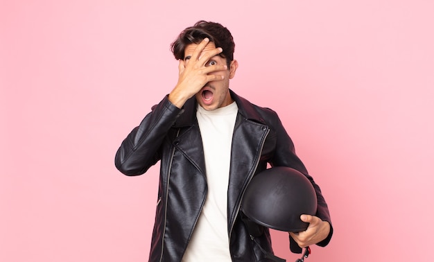 Hombre hispano joven que parece sorprendido, asustado o aterrorizado, cubriendo la cara con la mano. concepto de piloto de moto