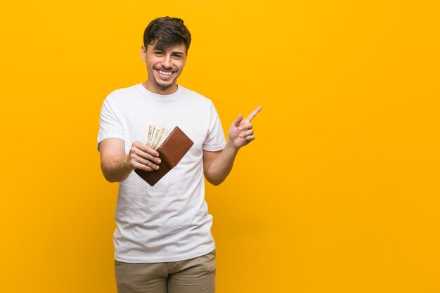 Foto hombre hispano joven que sostiene una billetera sonriendo alegremente apuntando con el dedo índice.