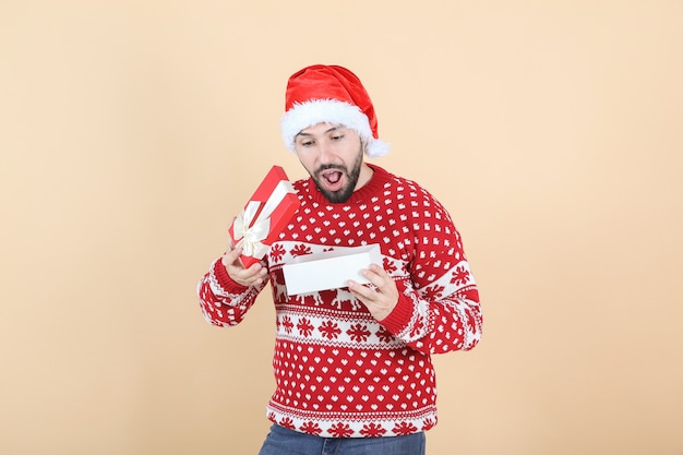 Hombre hispano latino, con sombrero de Navidad abriendo un regalo, fondo beige