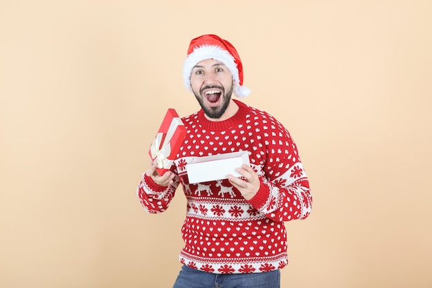 Hombre hispano latino, con sombrero de Navidad abriendo un regalo, fondo beige
