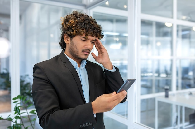 Hombre hispano molesto en la oficina en el trabajo leyendo malas noticias del hombre de negocios en línea del teléfono en camisa cerca