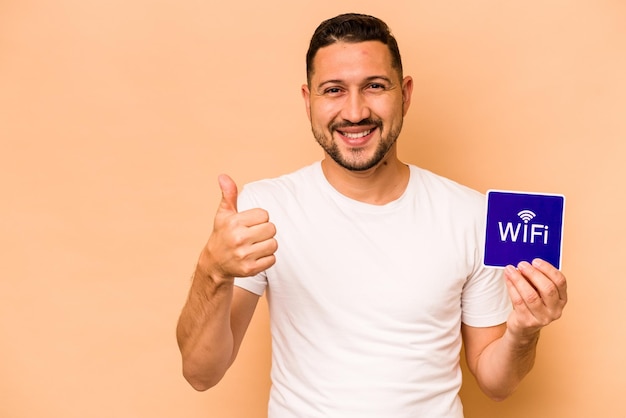 Hombre hispano sosteniendo un cartel wifi aislado de fondo beige sonriendo y levantando el pulgar