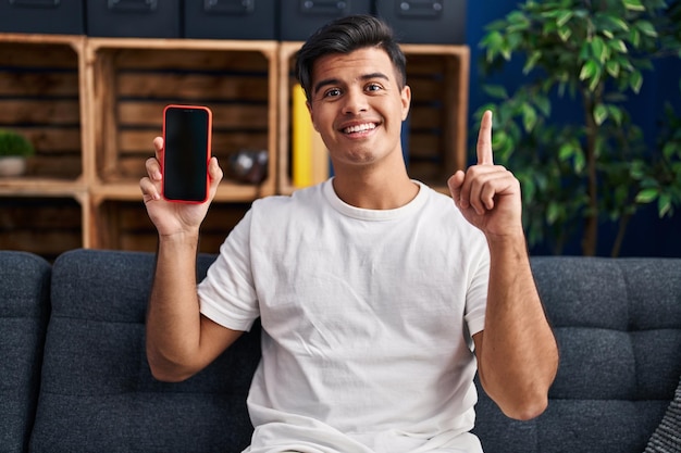 Hombre hispano sosteniendo un teléfono inteligente que muestra una pantalla en blanco sorprendido con una idea o pregunta señalando con el dedo con cara feliz, número uno