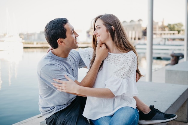 Un hombre hispano sostiene a su novia sonriente mientras ambos están sentados en el muelle en España
