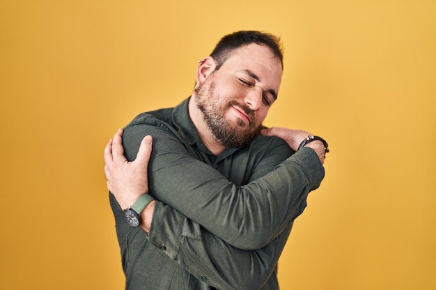 Hombre hispano de talla grande con barba parado sobre fondo amarillo abrazándose a sí mismo feliz y positivo sonriendo confiado amor propio y cuidado personal